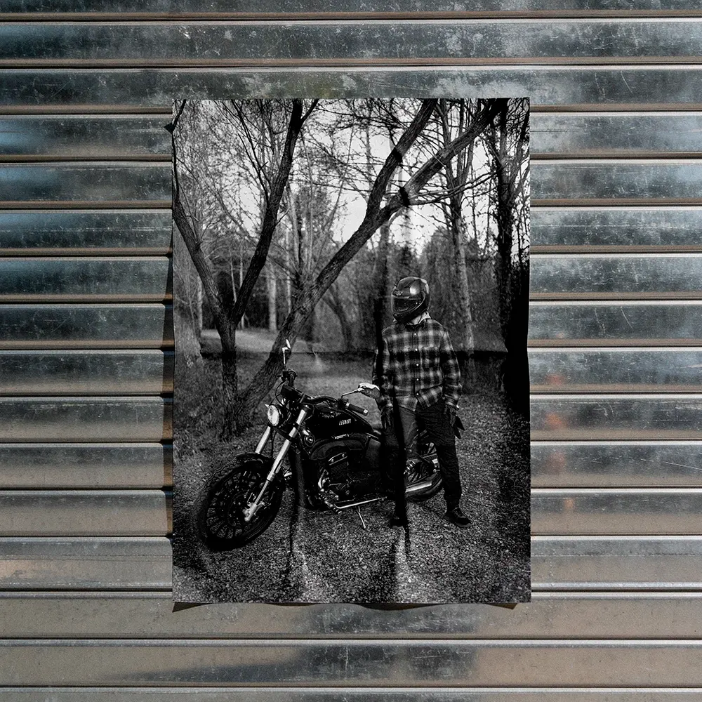 Black and white poster pasted on a corrugated metal shutter showing a helmeted rider beside a custom motorcycle in a bare forest