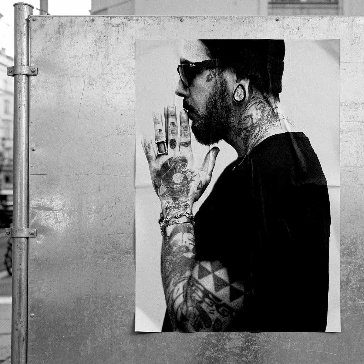 Black and white photographic poster pasted on an urban metal panel, side portrait of a tattooed man in a beanie and sunglasses, street photography in Bilbao
