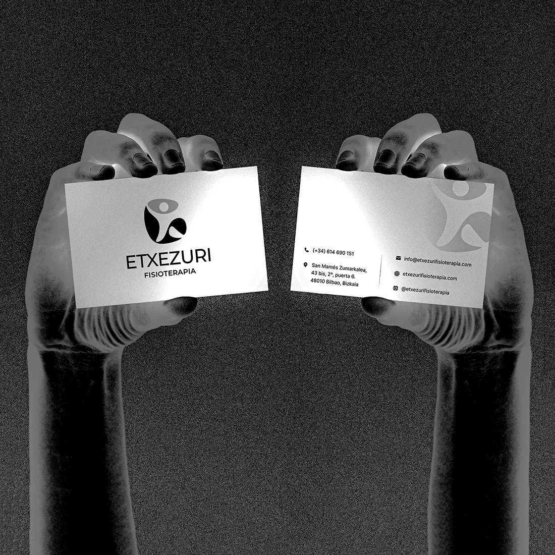 Two hands hold Etxezuri Fisioterapia business cards, the logo on the front and contact details on the back, in grainy black and white.