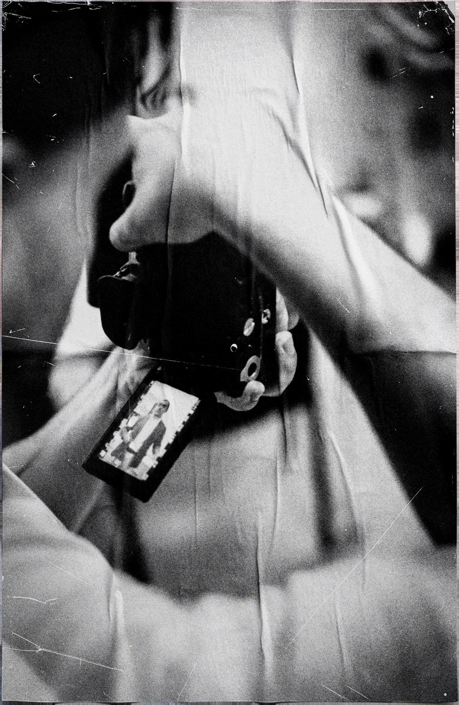 Black and white backstage detail of a shoot: hands hold a camera with a developed film frame hanging from it, over a crumpled-paper texture.