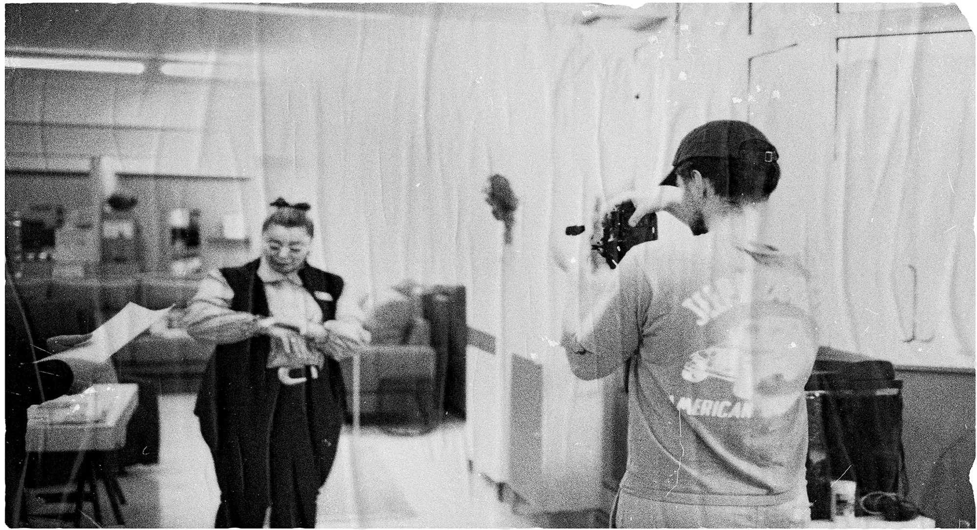 Black and white making of a studio shoot: an operator handheld-films a styled model beside a white backdrop, with a film-frame border.