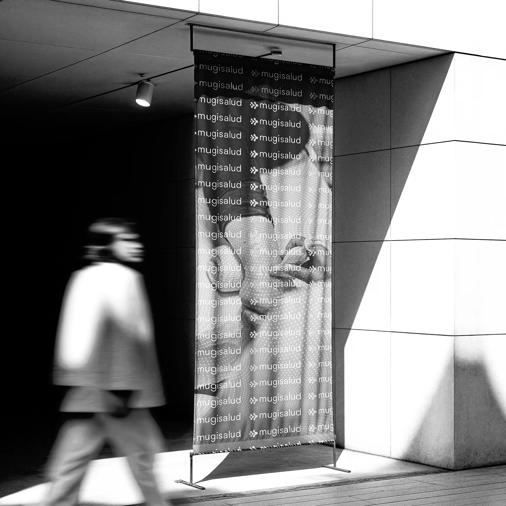 Vertical roll-up banner for Mugisalud at a modern building entrance, its white logo repeated over a black-and-white physiotherapy photo.