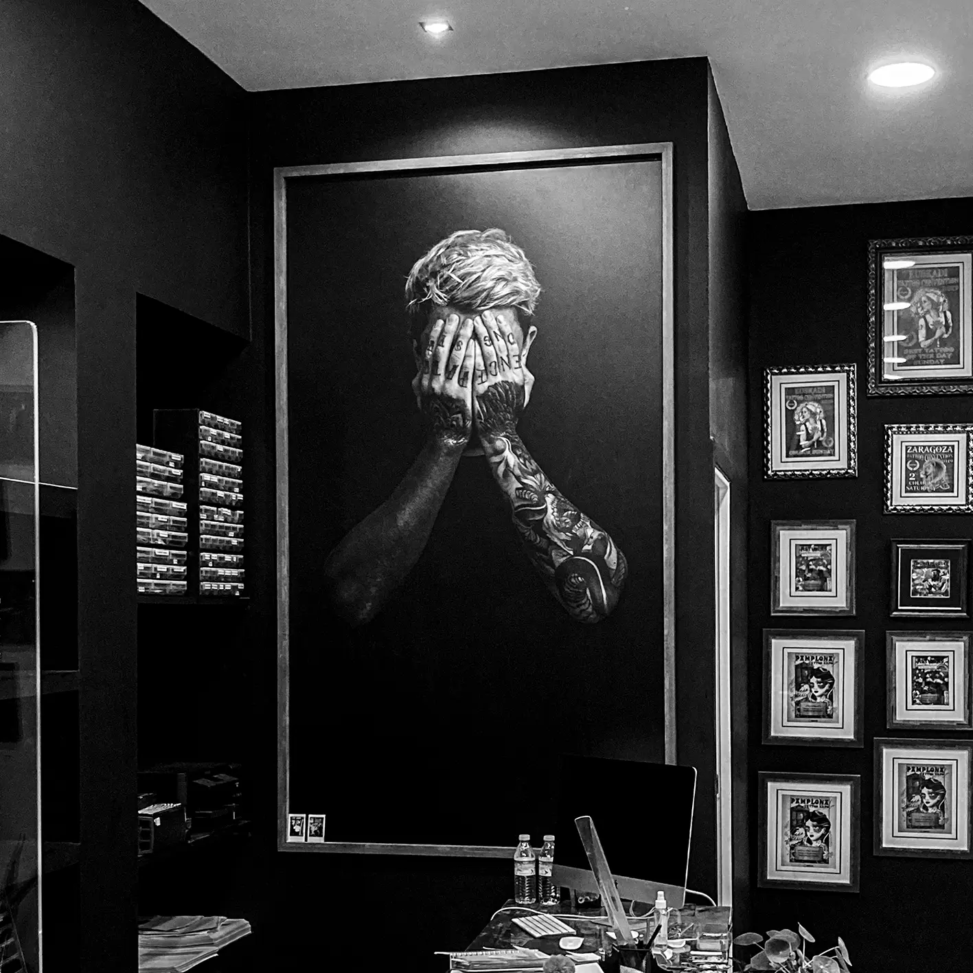 Black-and-white mural of a man covering his face with tattooed hands, in a Bilbao tattoo studio beside framed posters.