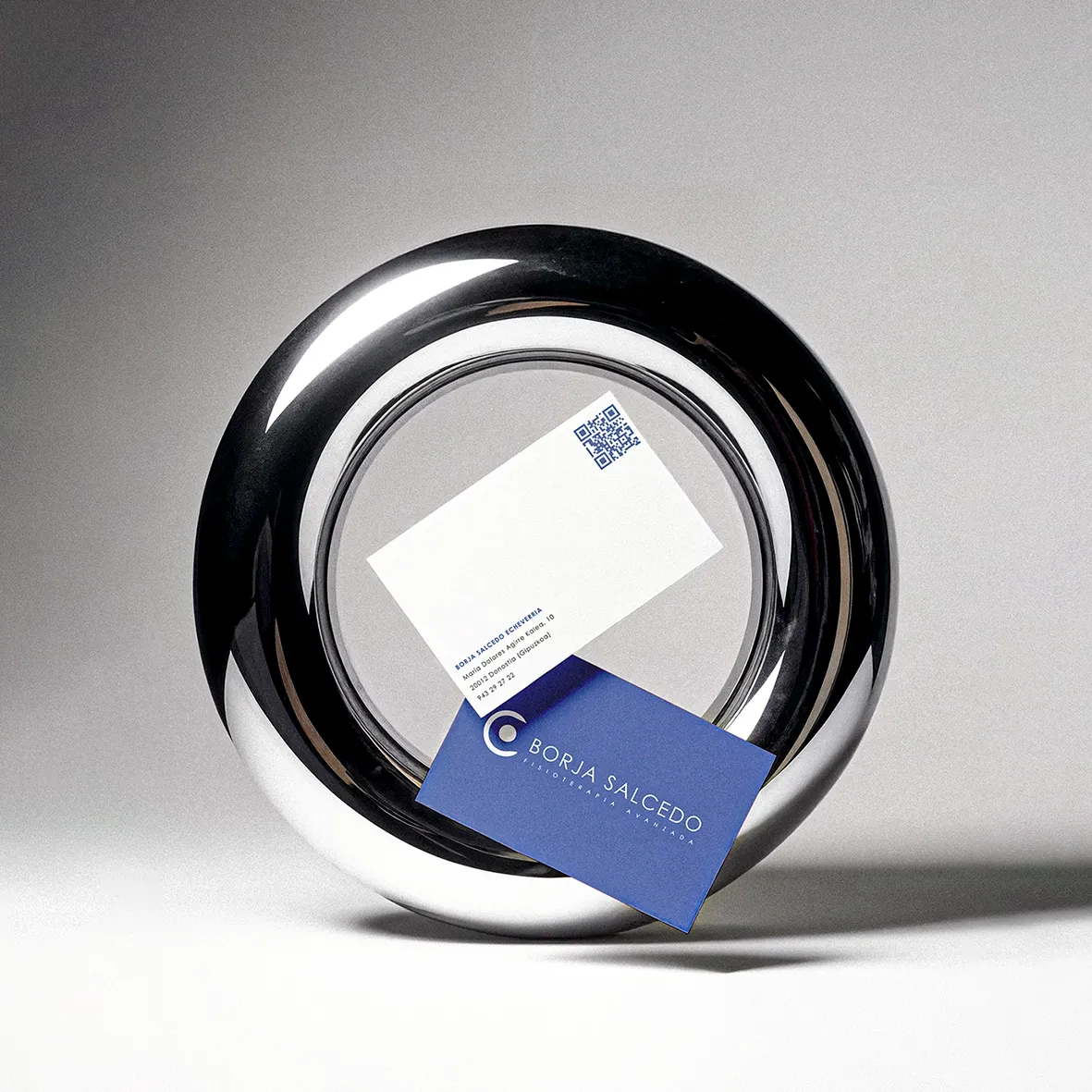 Business cards for Salcedo Fisioterapia Avanzada in blue and white, resting against a polished chrome ring on a grey backdrop